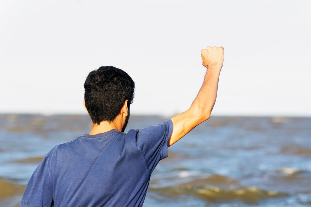 Man in blue T-shirt facing the ocean with fist raised in triumph